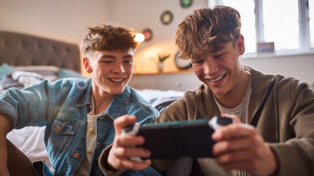 Dos jóvenes sonrientes sentados en una habitación mientras juegan juntos en una consola portátil.