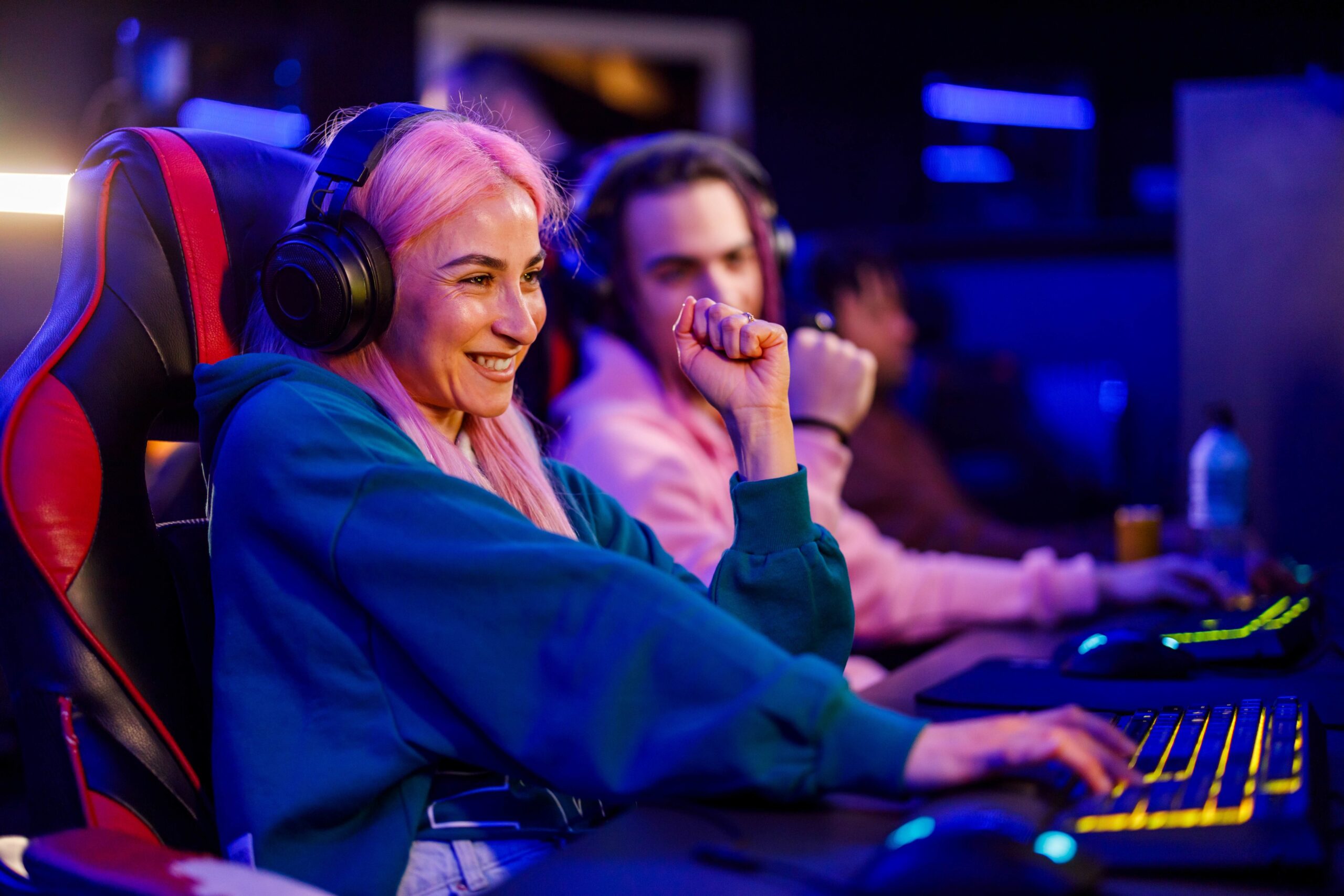 Mujer gamer con cabello rosa y auriculares, celebrando frente a teclado retroiluminado en competencia de videojuegos.